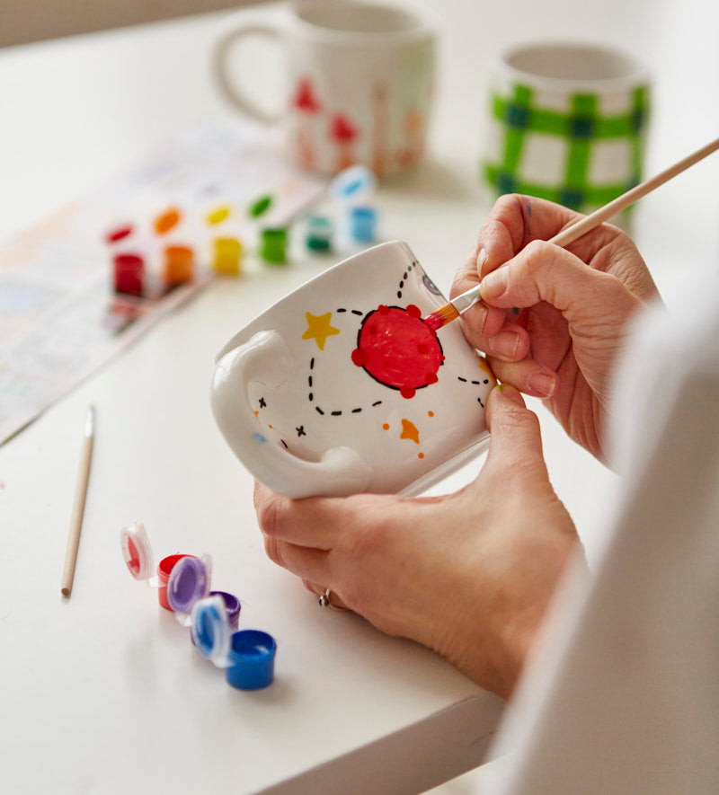 Stacked white mugs with colorful hand-painted designs including a rainbow, planets, and cherries on a light-colored surface.