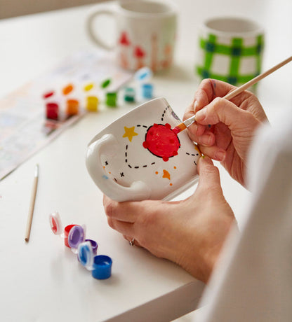 Stacked white mugs with colorful hand-painted designs including a rainbow, planets, and cherries on a light-colored surface.